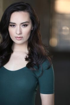 a woman with long dark hair wearing a green shirt and looking at the camera while standing in an empty room