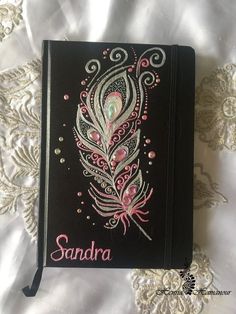 a black notebook with pink and white designs on it sitting on a lace covered surface