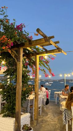 an outdoor dining area with tables and chairs near the water at dusk, along with flowers on the pergolated roof