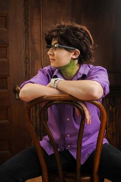 a woman sitting in a chair with her arms crossed, wearing glasses and a purple shirt