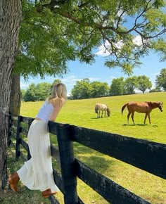 a woman leaning on a fence looking at horses grazing in the field behind her is a tree