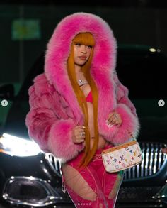 a woman wearing a pink fur coat and holding a white purse in front of a car