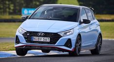 the front end of a blue car driving on a race track with grass and trees in the background