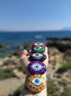 a person's hand holding five painted rocks in front of the ocean with an evil eye on it