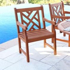 two wooden chairs sitting next to each other on a patio near a pool with water in the background