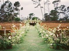 an aisle lined with rows of wooden benches covered in white flowers and greenery next to trees