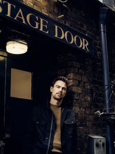 a man standing in front of a brick building wearing a black jacket and tan sweater