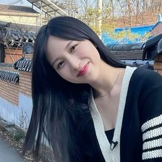 a woman with long black hair standing next to a brick wall and looking at the camera