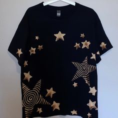 a black t - shirt with gold stars and spirals on the front is hanging against a white wall