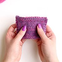 two hands holding up a small purple bag on top of a white table next to a pink heart