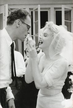 a man standing next to a woman in a wedding dress and holding a piece of cake