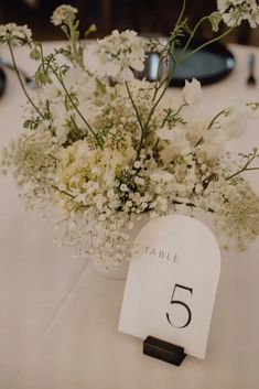 a table number with flowers in a vase on top of a white table cloth covered table