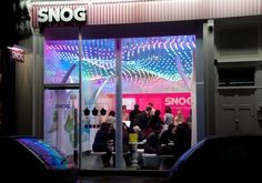 people sitting at tables in front of a store window with neon lights on the windows