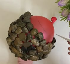 a hand holding a red ball with rocks and stones on it, next to purple flowers