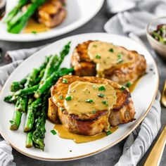 two white plates topped with meat covered in gravy next to asparagus