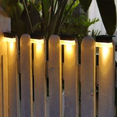 a white picket fence with lights on it and some plants in the backround