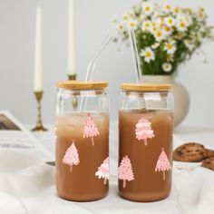 two mason jars filled with brown liquid and pink trees on the lids, sitting on a table next to cookies