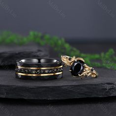 two wedding bands with black stones and gold accents on top of a stone slab, surrounded by greenery