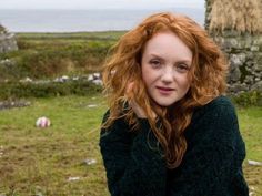 a woman with red hair and green sweater posing for the camera in front of some rocks