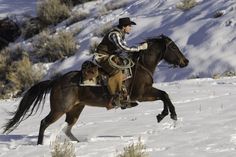 a man riding on the back of a brown horse in snow covered field next to shrubbery