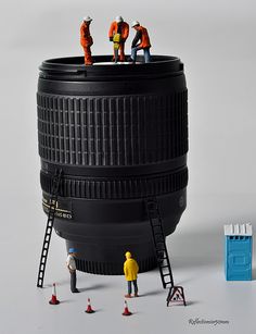 miniature people standing on top of a large camera lens with ladders to reach it