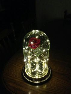 a lighted glass dome with a red rose in it sitting on a wooden table top