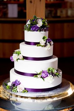 a three tiered cake with purple and white flowers on the top is sitting on a table