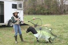 a woman holding a fishing rod next to a fake fish and a horse in front of a trailer