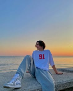 a young man sitting on the edge of a wall next to the ocean at sunset