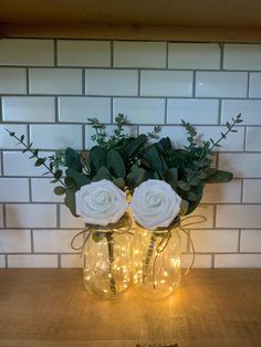 two vases with white roses and greenery are on a table in front of a brick wall