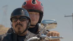 two people on a motorcycle with helmets and goggles, one riding the bike while the other is looking at the camera