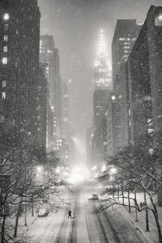 a black and white photo of a city street at night with snow falling on the ground