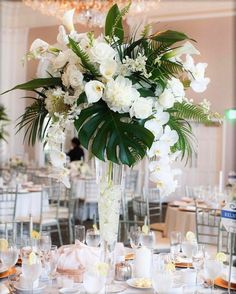 a tall vase filled with white flowers and greenery on top of a wooden table