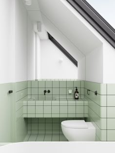 a white toilet sitting next to a bathroom sink under a slanted window in a green tiled wall