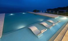 an empty swimming pool at night with lounge chairs on the edge and lights in the water