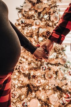 a pregnant woman and man holding hands in front of a christmas tree with ornaments on it