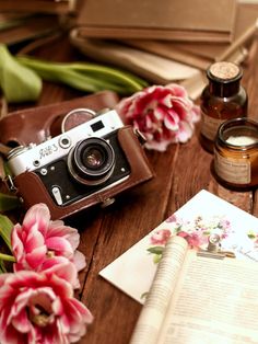 an open book, camera, and flowers on a wooden table with some writing material