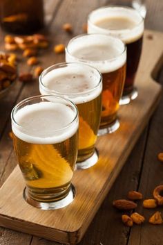 four glasses filled with beer sitting on top of a wooden table next to nuts and pretzels