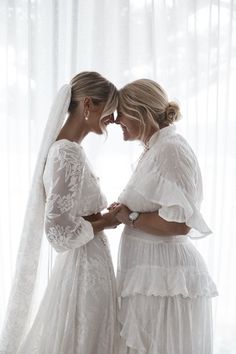 two women standing next to each other in front of a window wearing wedding gowns