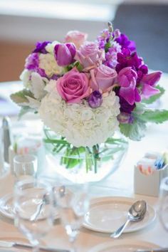 purple and white flowers in a glass vase on a table set for an elegant dinner