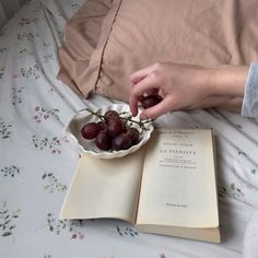 a person reaching for some grapes in a bowl on a bed with an open book