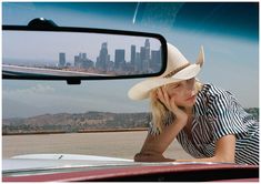 a woman wearing a cowboy hat leaning on the hood of a car in front of a rear view mirror