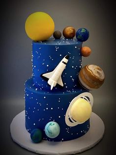 a three tiered blue cake with planets and rockets on the top, in front of a gray background