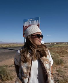 a woman wearing sunglasses and a hat standing in front of a welcome to arizona sign