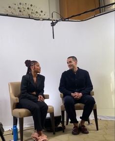 two people sitting on chairs in front of a white wall and one person standing up