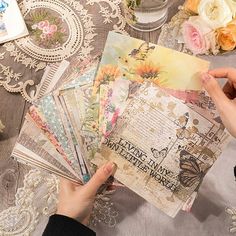 a person is holding up some papers on a table with flowers and other things in the background