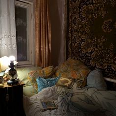 an unmade bed in front of a window with curtains on the side and a lamp next to it