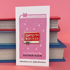 a pink enamel pin with the words sorry i'm booked on it sitting next to some books