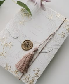 a wedding card with a wax stamp and tassel on it, sitting next to a flower