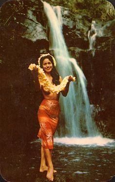 a woman standing in front of a waterfall wearing a hula skirt and holding flowers